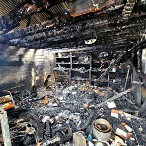 The burnt inside of a building, with charred roof timbers, a brick wall and shelves of burnt items