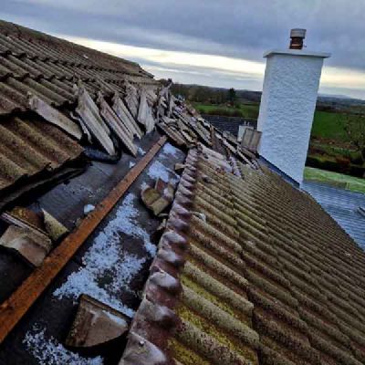 A roof of moss-covered tiles, with broken tiles and dark felt showing, beside a white chimney