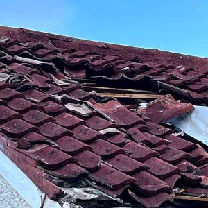 A roof of dark red tiles with a section missing and white sheeting showing